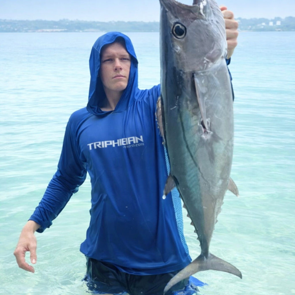 A person wearing a blue hooded shirt stands in clear water, holding up a large, freshly caught tuna by its gills.