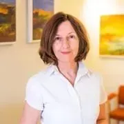 Kvinde i hvid skjorte smiler, stående i galleri med malerier på væggen.
