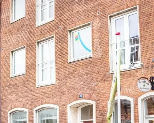 Murstensbygning med hvidindrammede vinduer; et med et farverigt logo. En vinduesvasker rækker et værktøj frem.
