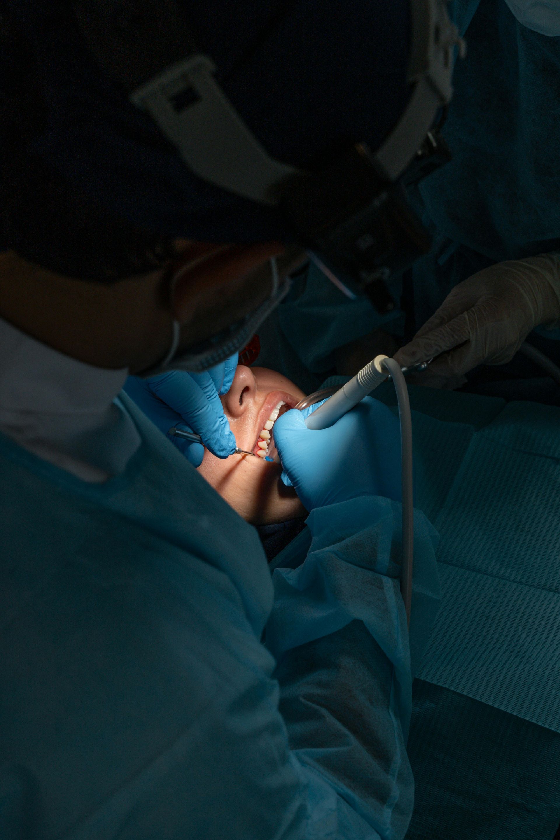 Tandlæge udfører procedure på patients åbne mund; patient i blåt kirurgisk draperi.