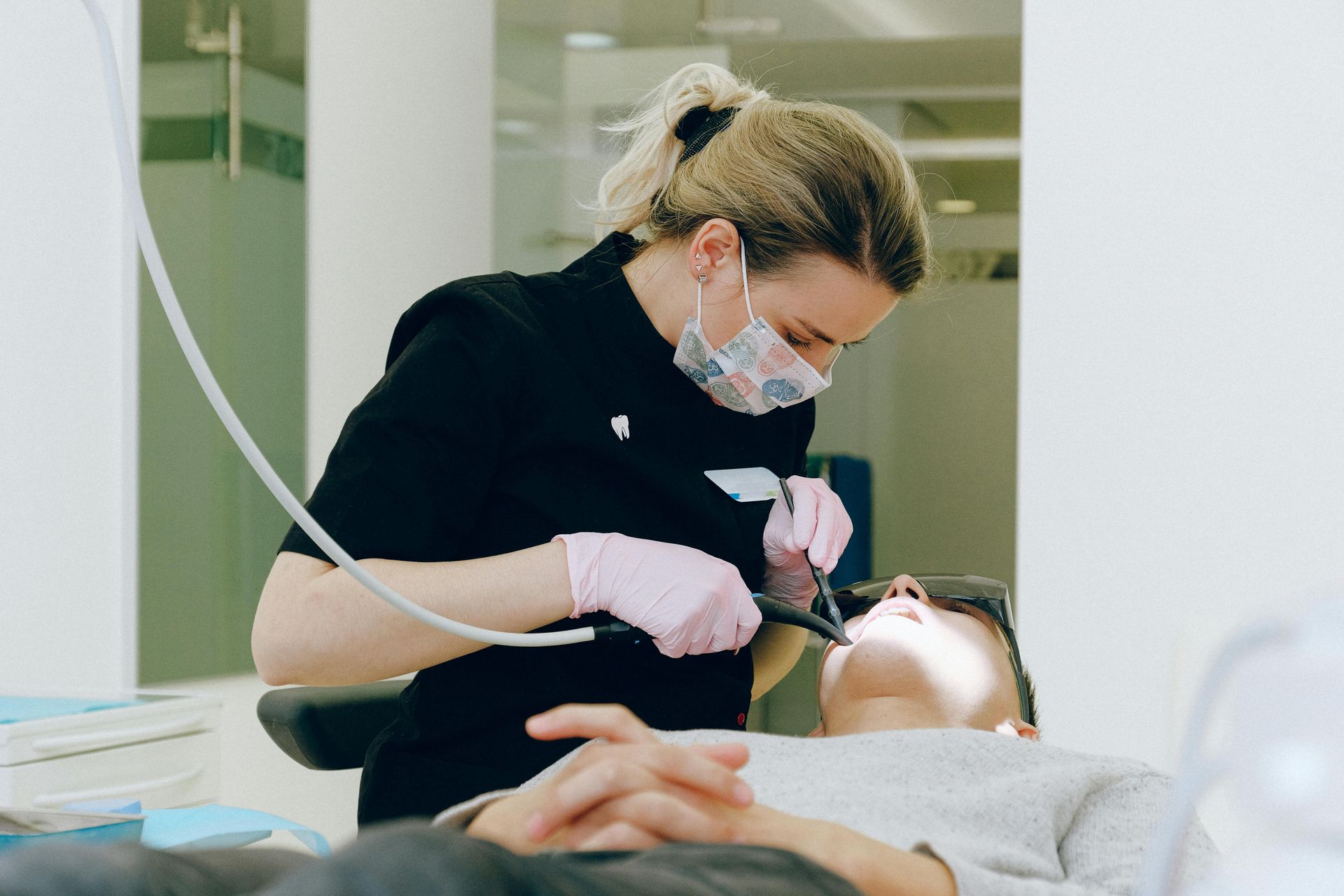 Tandlæge i maske, handsker og sorte scrubs arbejder på en patient i en tandlægestol.