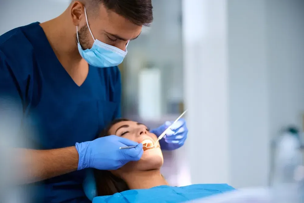 Tandlæge undersøger en patients tænder på en tandklinik. Begge har masker og handsker på.