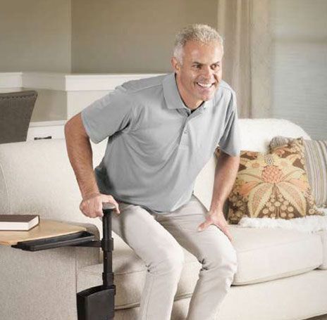 A person uses a sofa-mounted standing assist handle with a side tray to stand up from a beige couch in a living room.