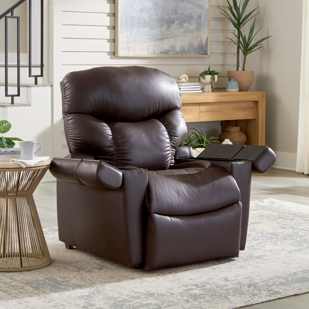 A dark brown leather recliner sits on a patterned area rug in a brightly lit living room near a wooden table and stairs.