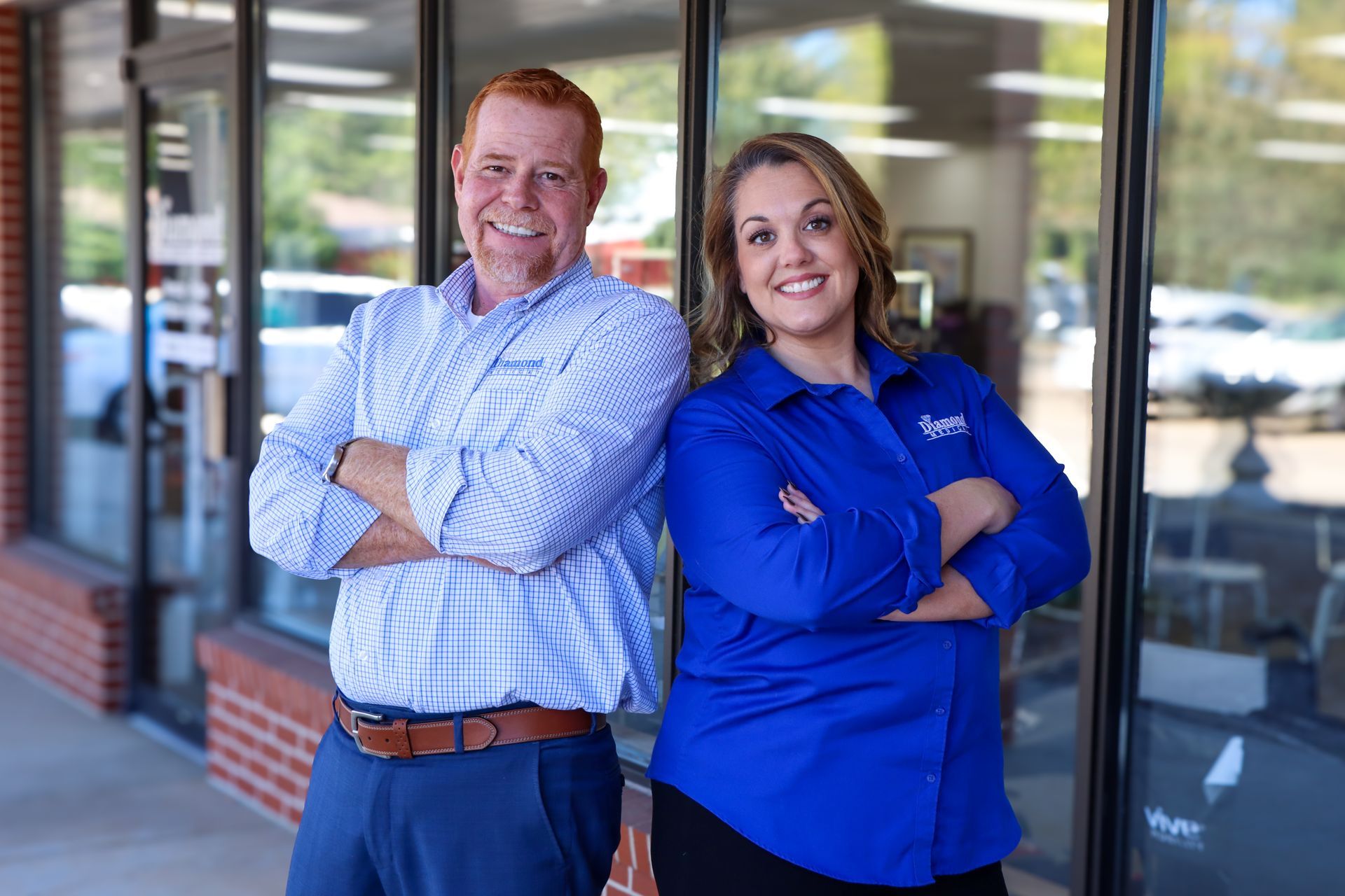 Two smiling people in professional attire stand with their arms crossed in front of a glass storefront.