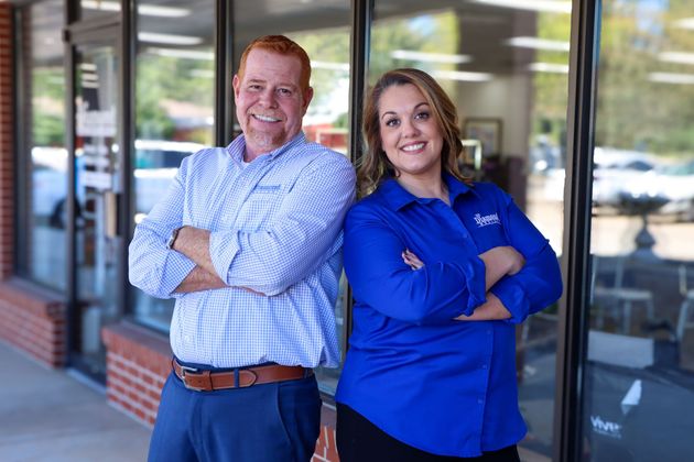 Two smiling people in professional attire stand with their arms crossed in front of a glass storefront.