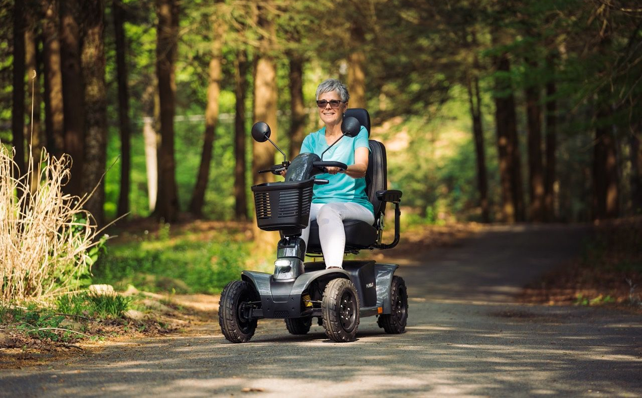 A person riding a mobility scooter on a sunlit, paved path through a forest.