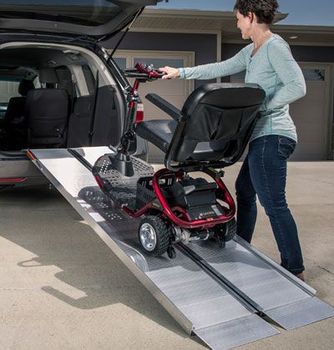 A person uses an aluminum ramp to load a red mobility scooter into the back of a parked SUV.