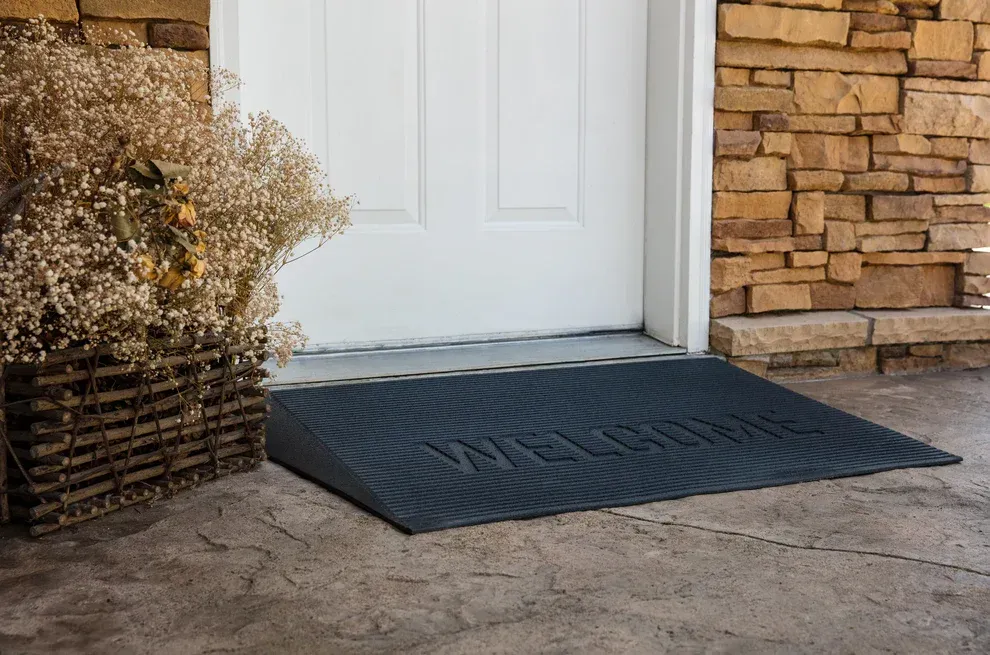 A black ramped welcome mat sits on a porch in front of a white door next to a stone wall and a rustic basket of flowers.