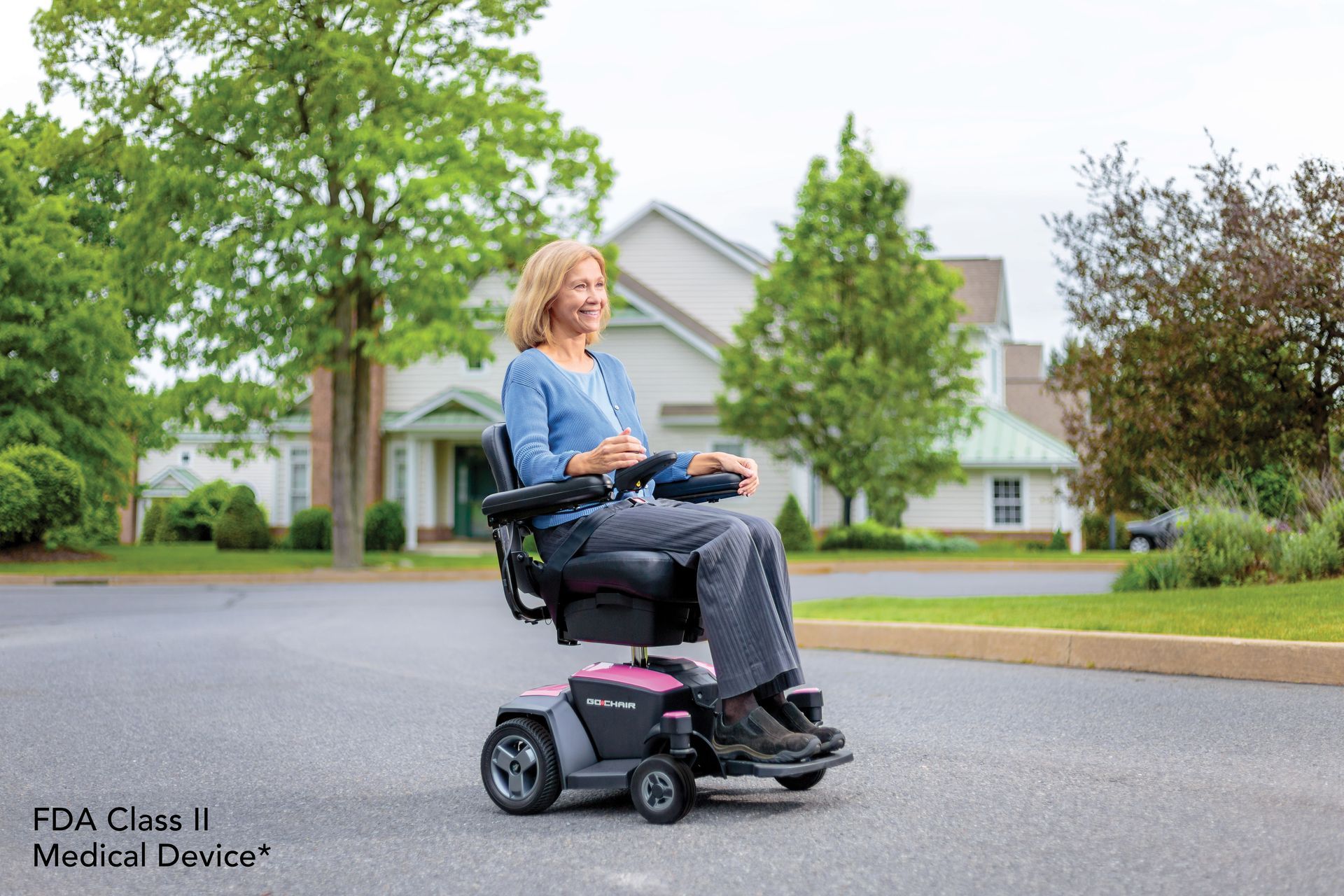 A person smiles while driving a dark gray and pink motorized wheelchair on an asphalt path in front of a house.