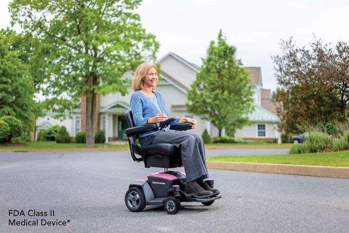 A person smiles while driving a dark gray and pink motorized wheelchair on an asphalt path in front of a house.