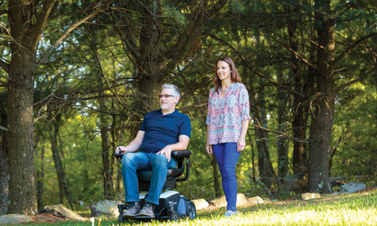A person in a motorized wheelchair and a companion walk together along a grassy path in a sunlit, wooded area.