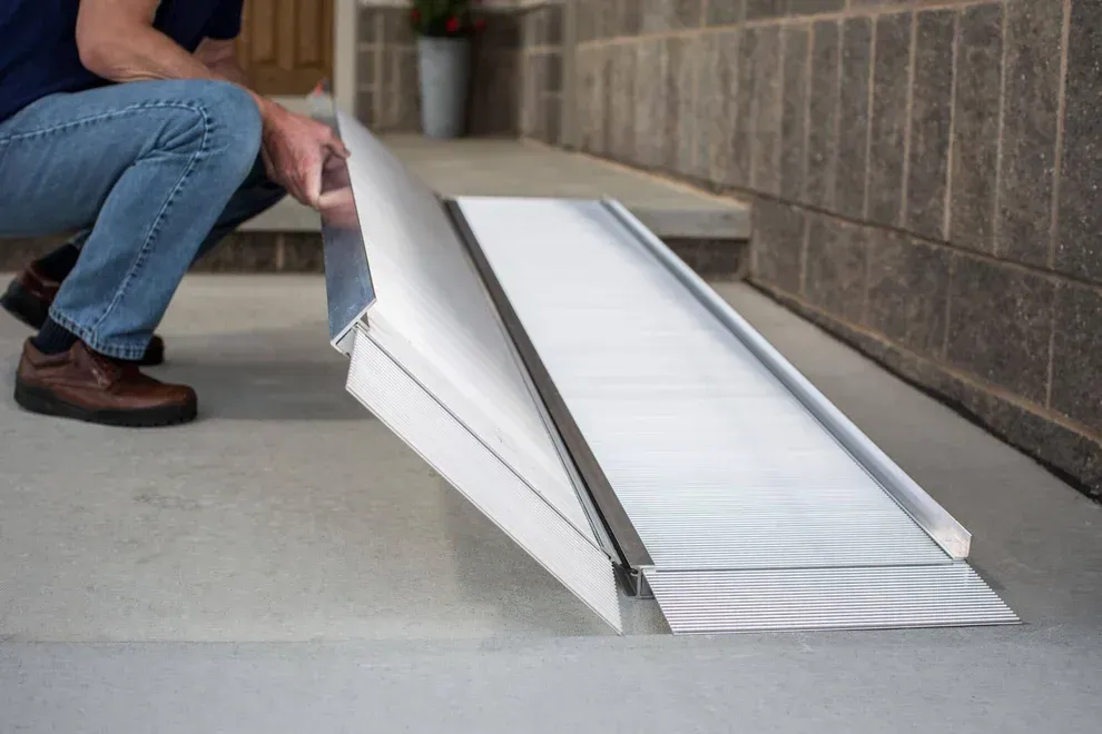 A person sets up a foldable aluminum wheelchair ramp on a concrete step leading to a building entrance.