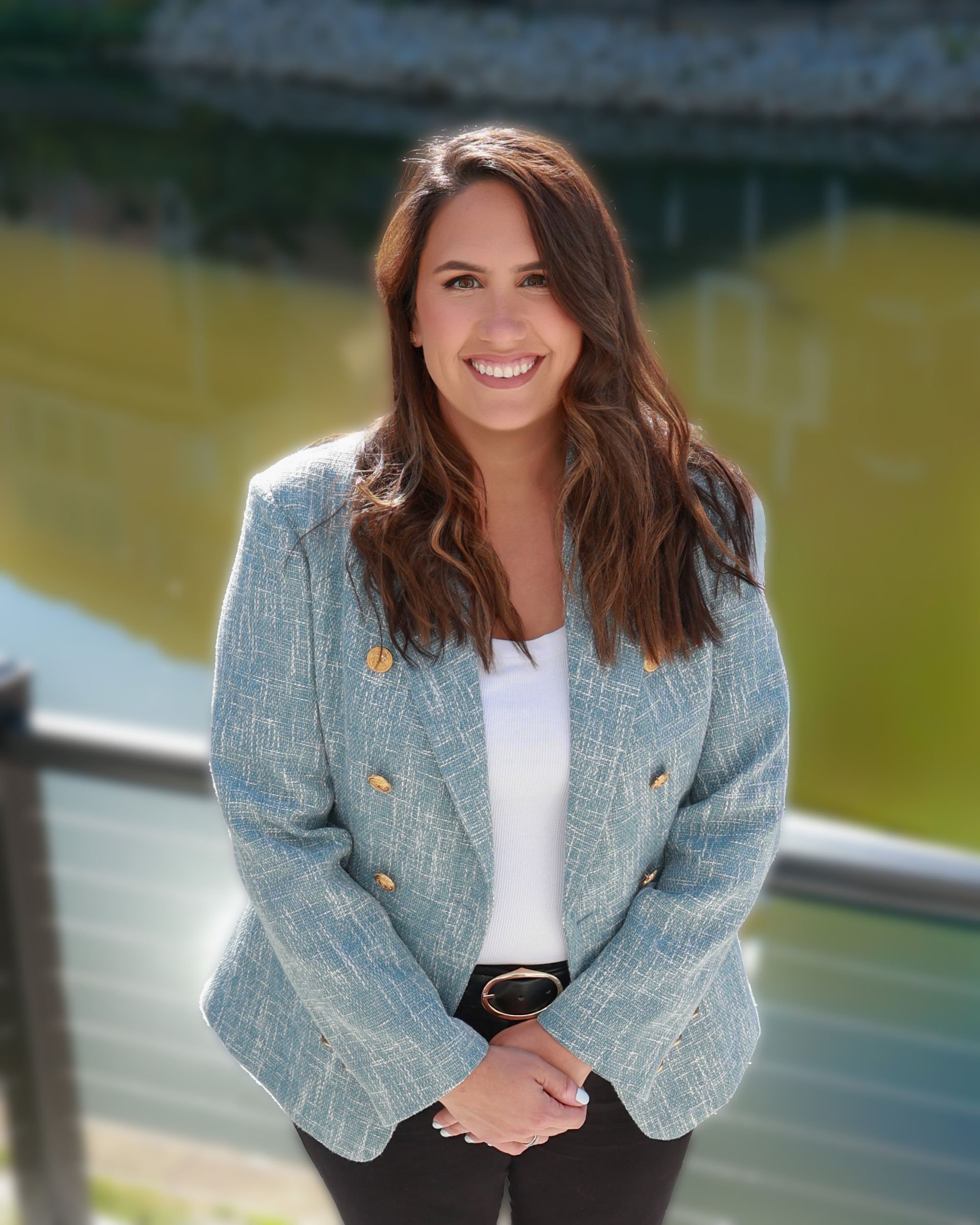 Smiling woman in a light blue blazer standing outdoors near water.