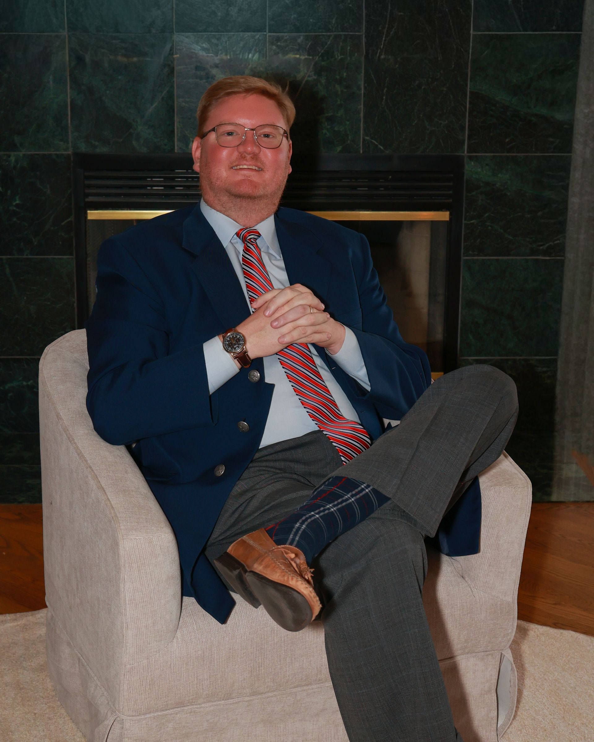Man in a blue blazer sits cross-legged in a light armchair before a dark tiled fireplace.