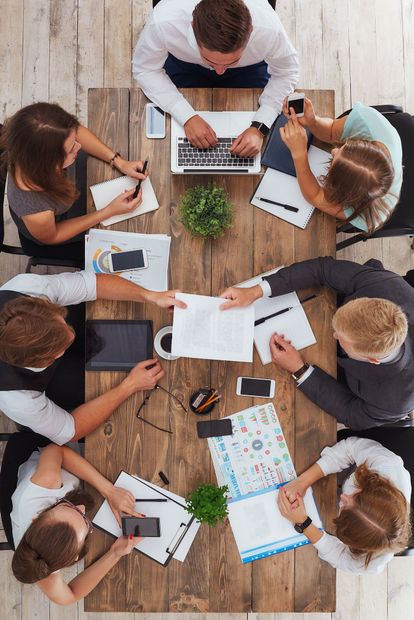 People in a business meeting around a wooden table, working on laptops, papers, and a tablet.