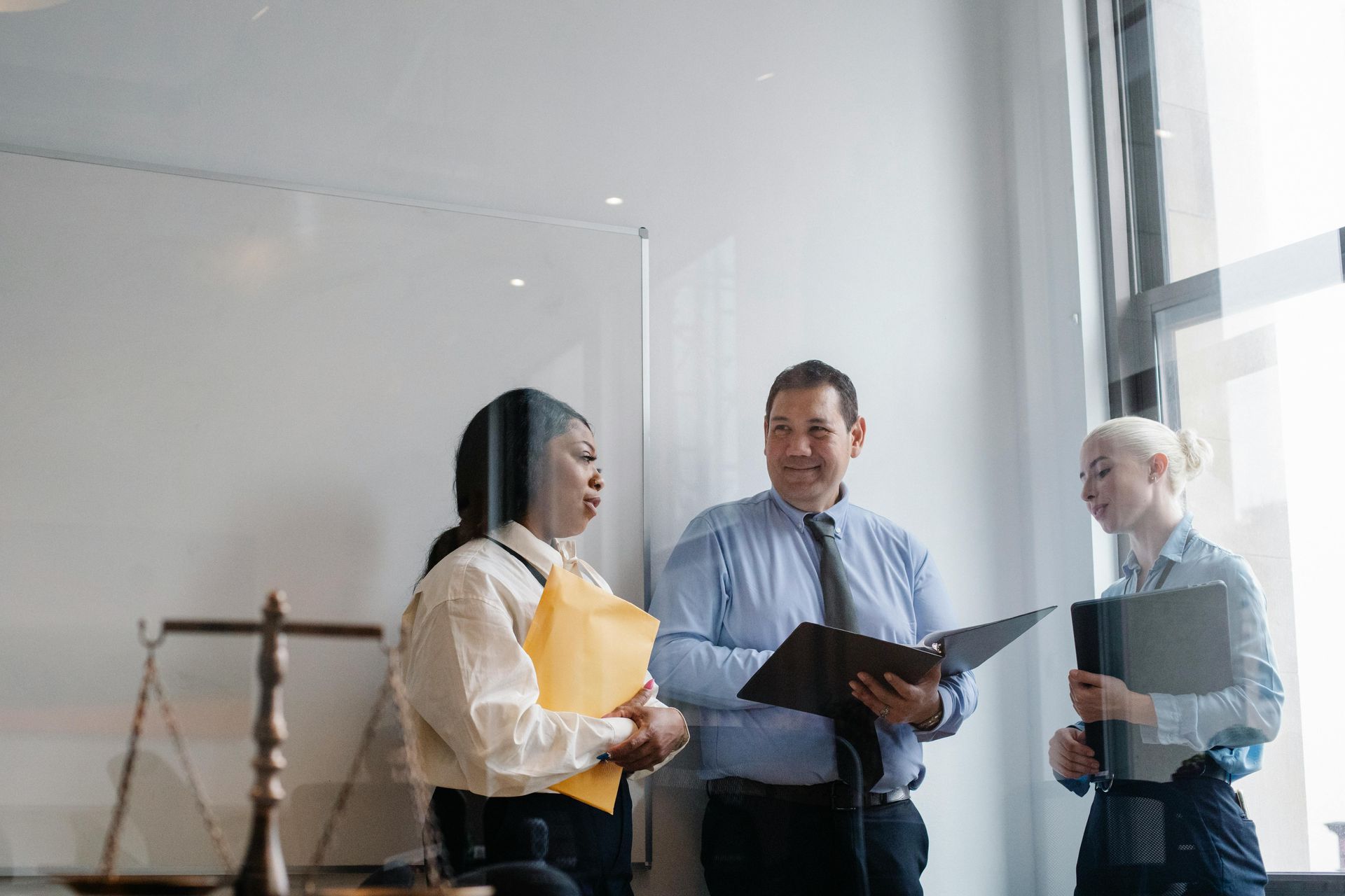 Three coworkers in an office hallway discussing documents near a whiteboard