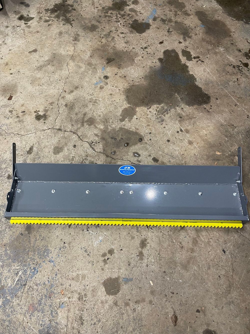 Gray metal snow plow blade with yellow rubber edge, on a concrete surface.
