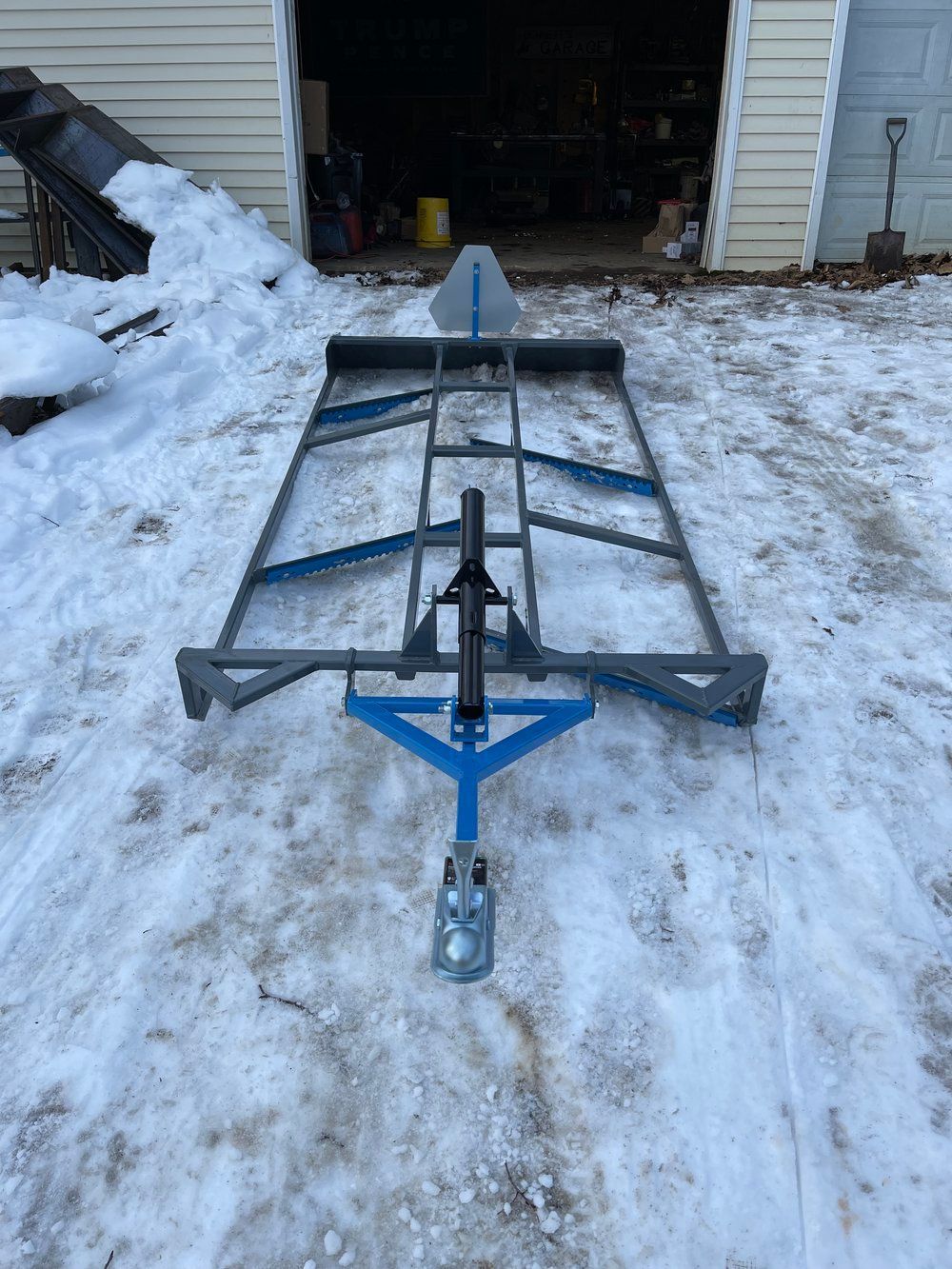 A blue trailer is sitting in the snow in front of a garage.