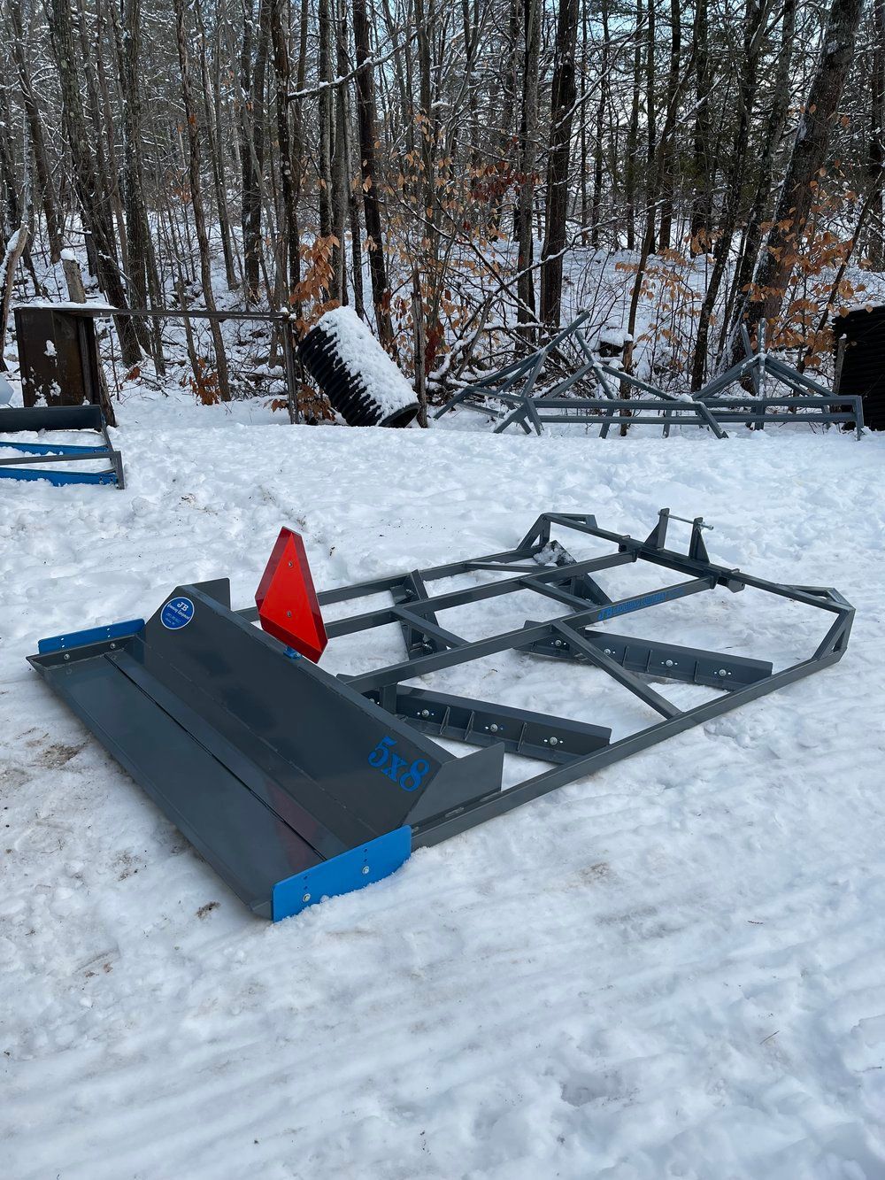 A grey tractor blade on snow, with a red triangle, set in a snowy forest.
