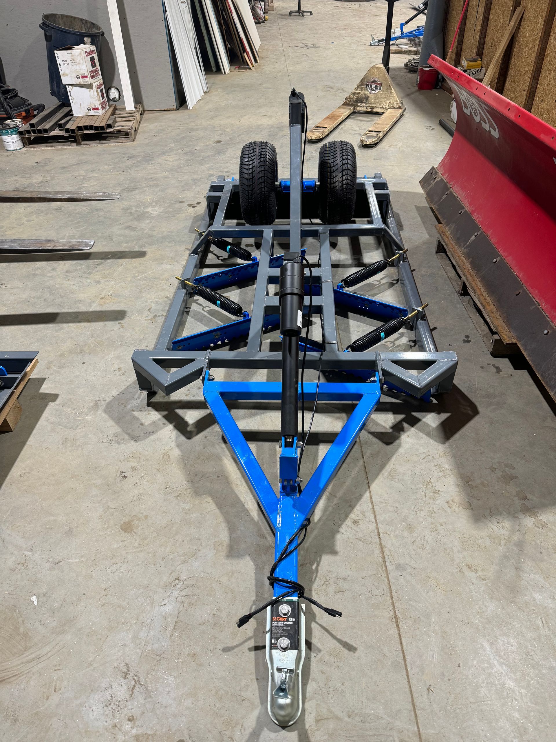 A blue trailer is parked in a warehouse next to a red plow.