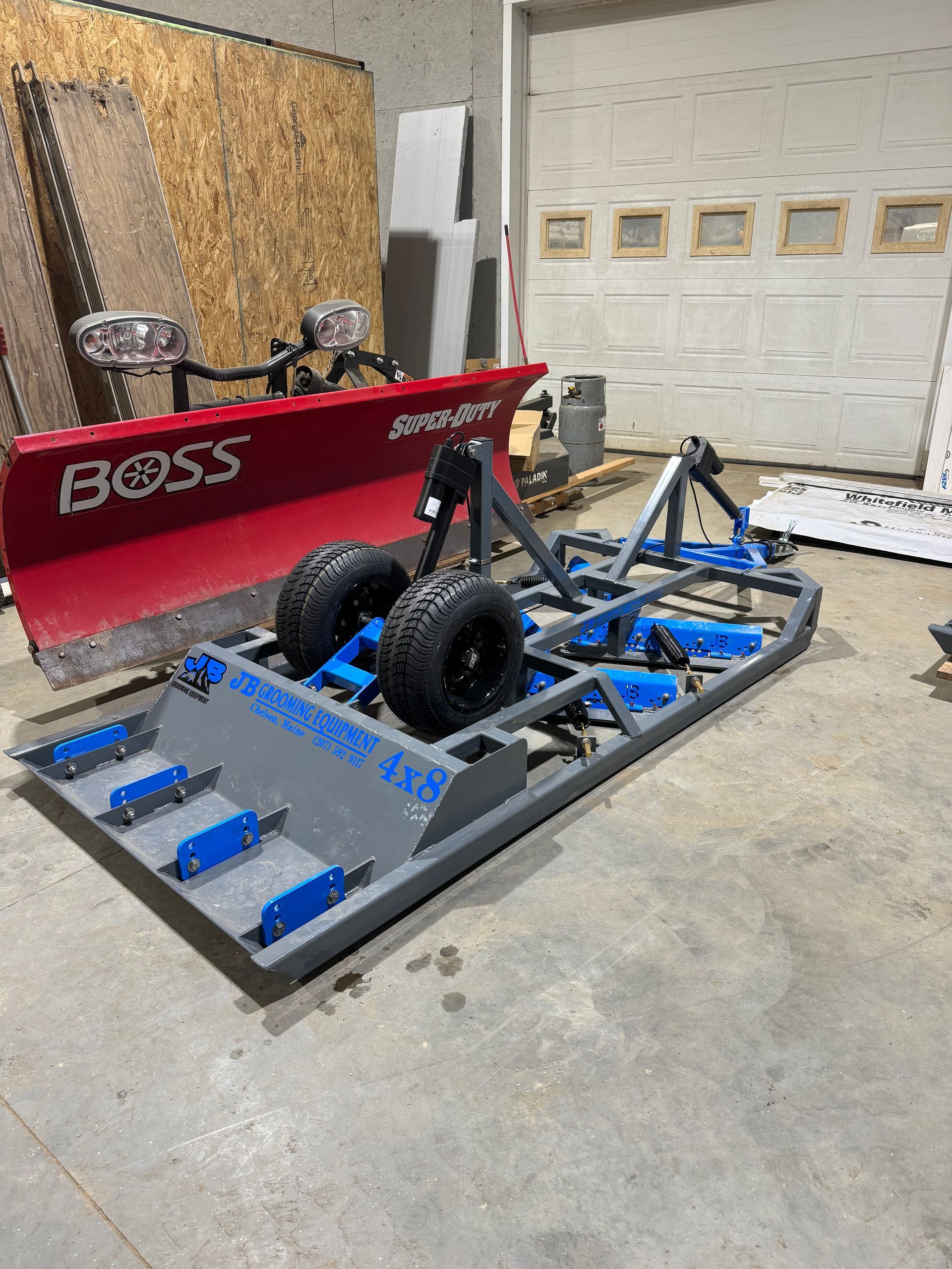 A gray and blue grading and leveling tool with two wheels and a red snowplow in a garage.