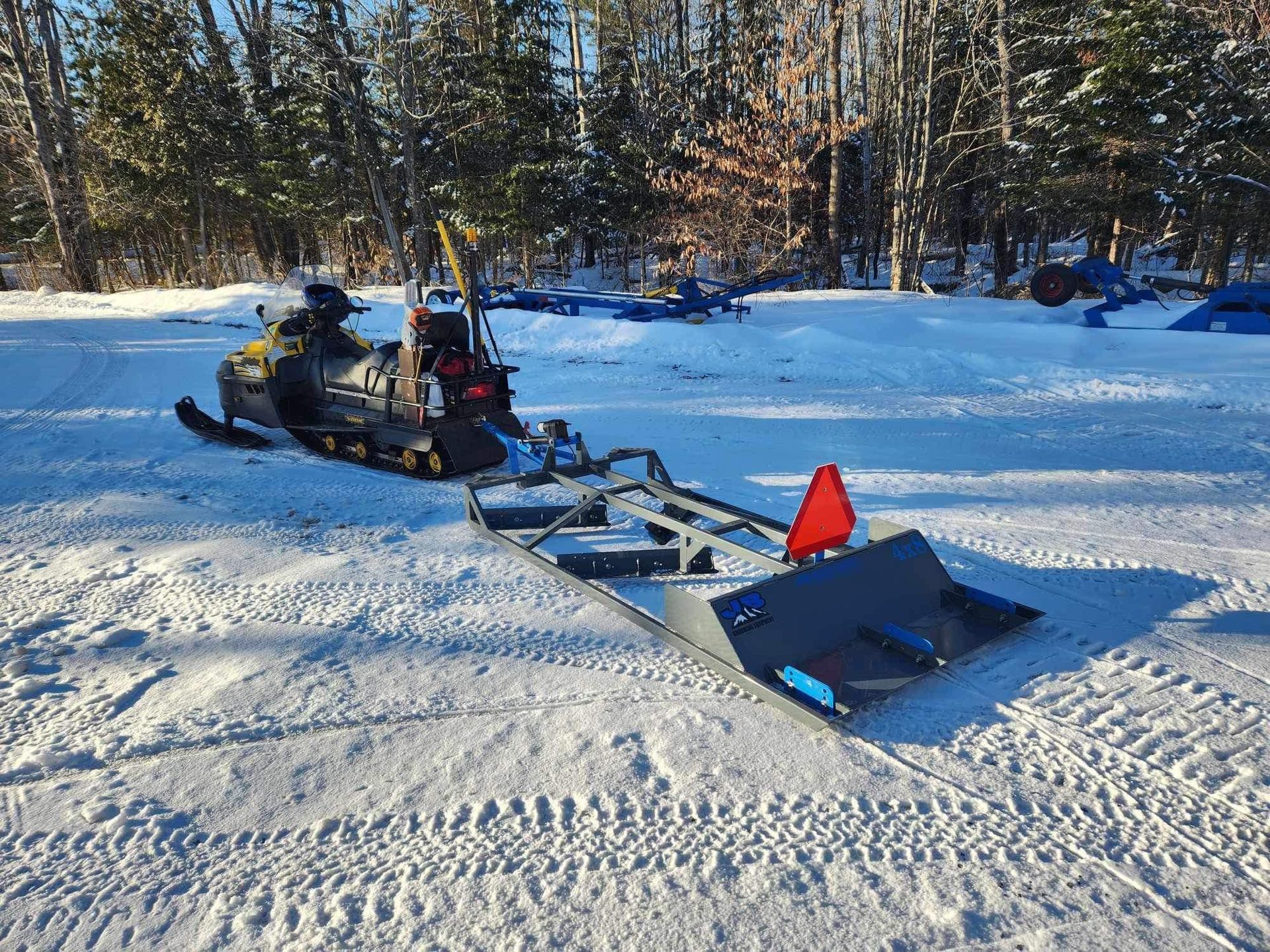 Snowmobile pulling a snow plow on a snowy path with trees in the background. Orange triangle on plow.