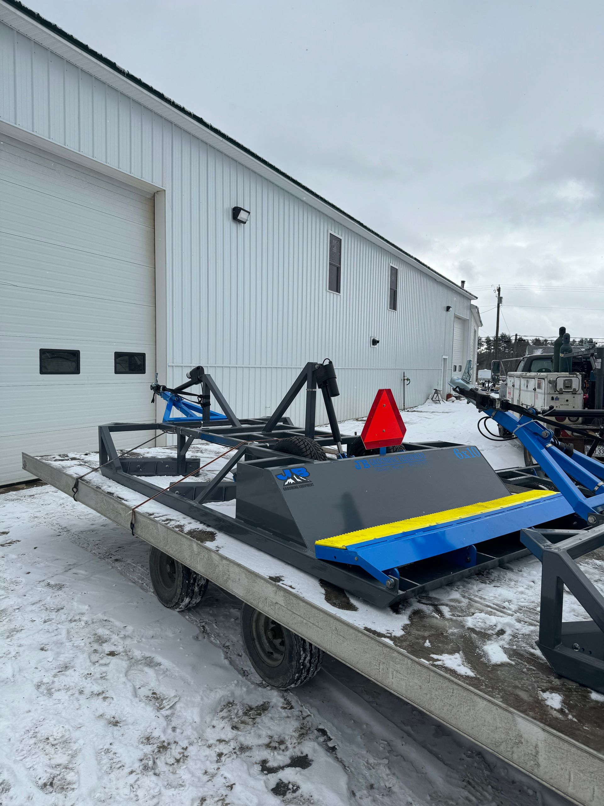 A blue and yellow tractor is parked in the snow in front of a white building.
