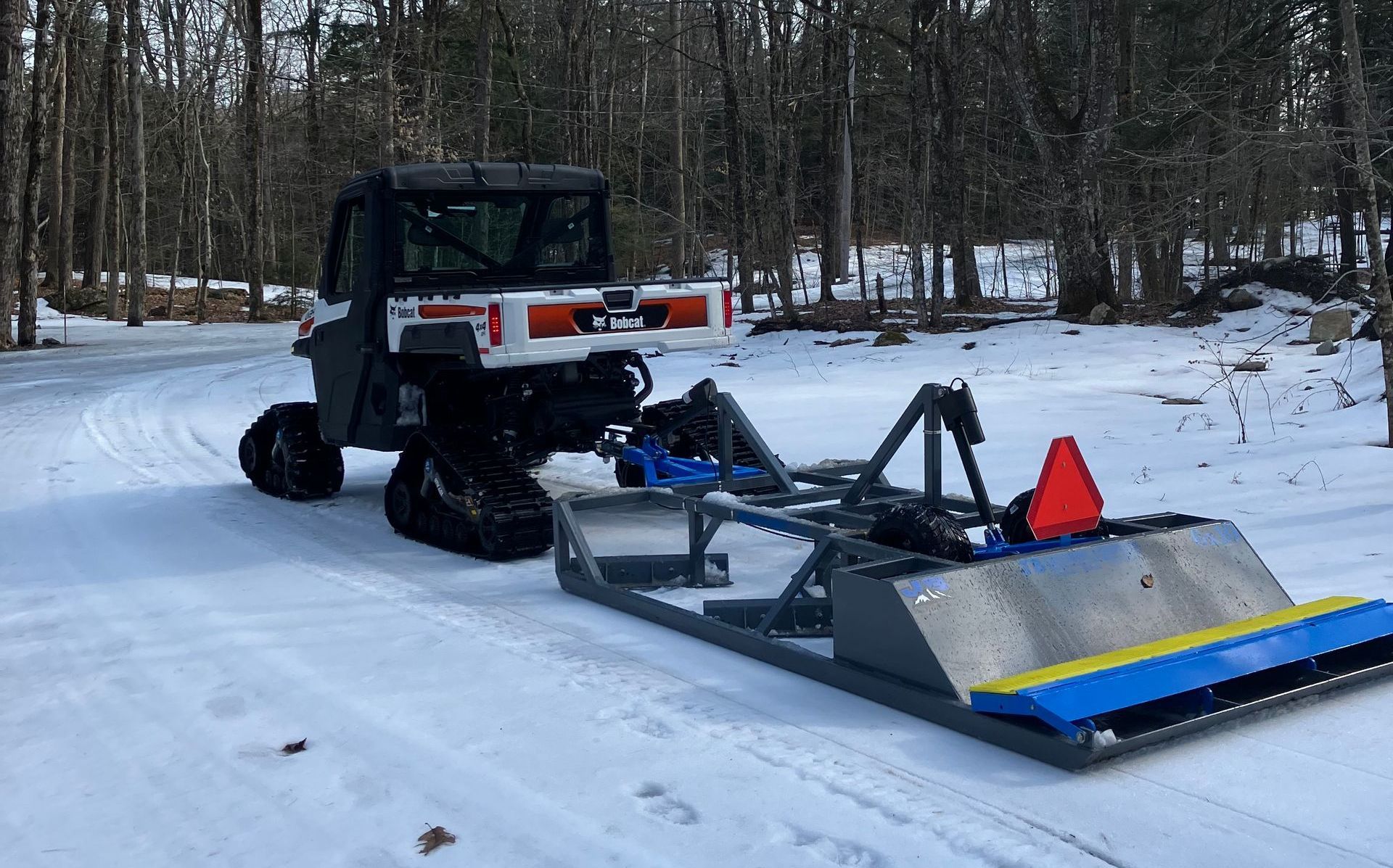 Bobcat utility vehicle with tracks pulling a snow groomer on a snow-covered path.