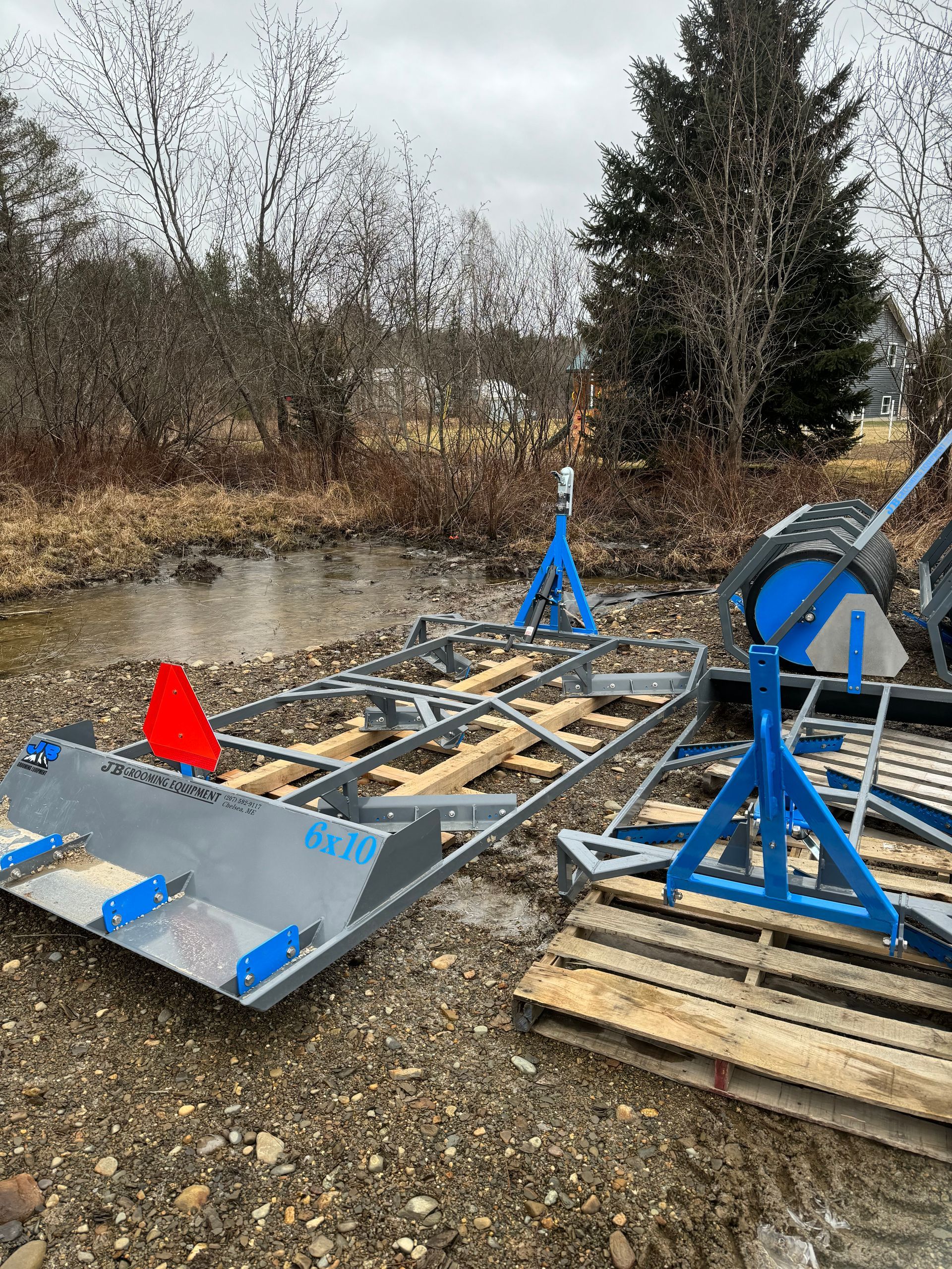 A large, metal and wooden land leveler sits in a wet, outdoor area, near water and some pallets.