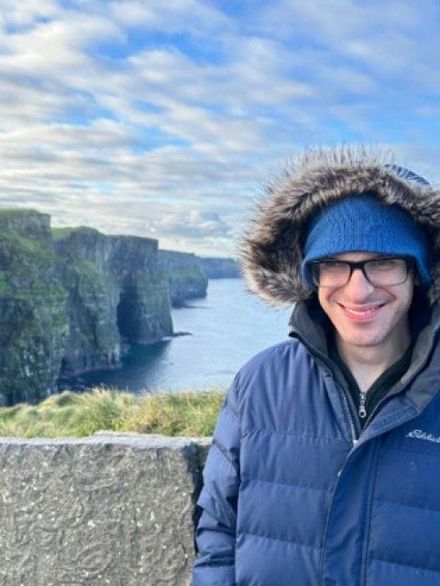 Person in blue jacket and fur hood standing by sea cliffs under a cloudy sky.