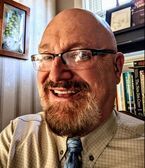 Smiling bald man with glasses and goatee in a tie, seated in a book-lined office