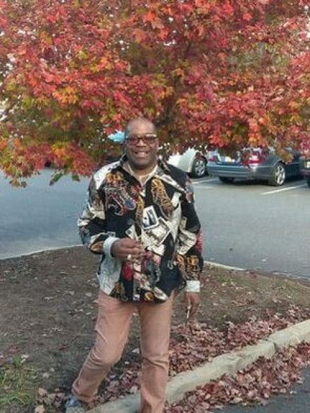 Person standing outdoors by a tree with red leaves, smiling and holding a drink, with parked cars behind.