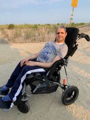 Man reclining in a black wheelchair on a sandy beach near dunes and the shoreline.