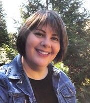 Smiling person in a denim jacket outdoors with evergreen trees in the background