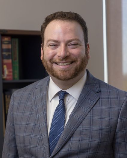 A man in a white shirt and tie is smiling in front of a bookshelf filled with books.