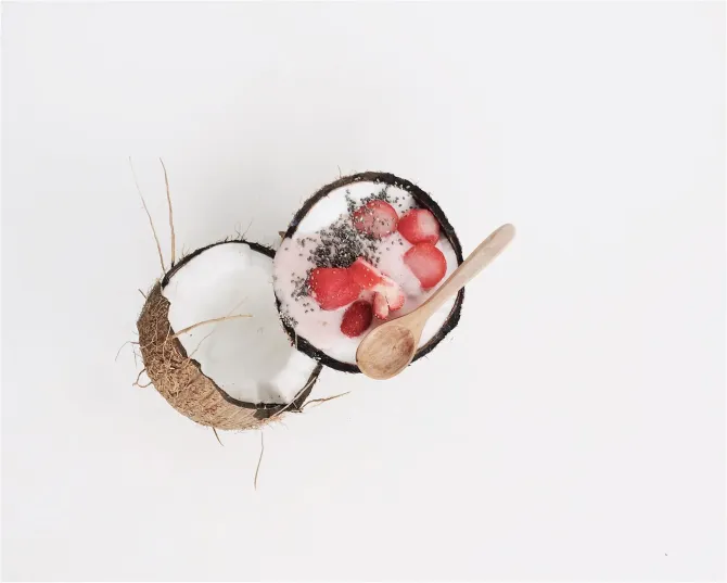 Coconut shell with a pink smoothie, strawberries, and a wooden spoon on a white background.