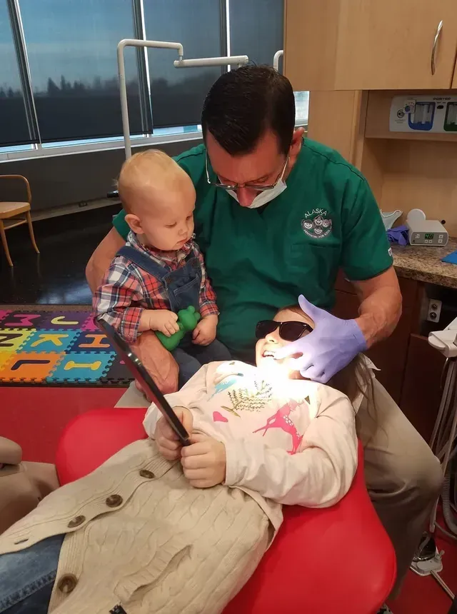 Dentist examining a child's teeth, while holding a toddler. Both children are looking at the dentist.