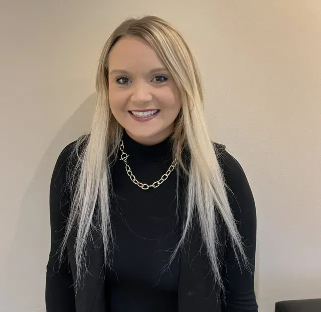 Woman with long blonde hair smiles, wearing a black turtleneck and vest, silver chain. Neutral background.