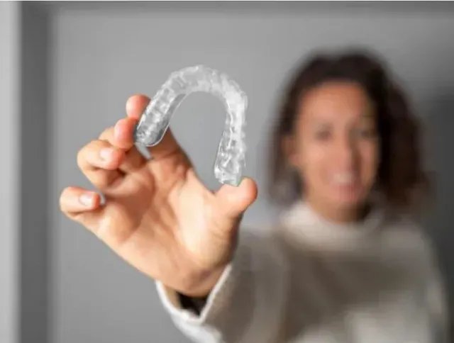 Woman holding a clear, plastic dental aligner, smiling, against a blurred gray background.