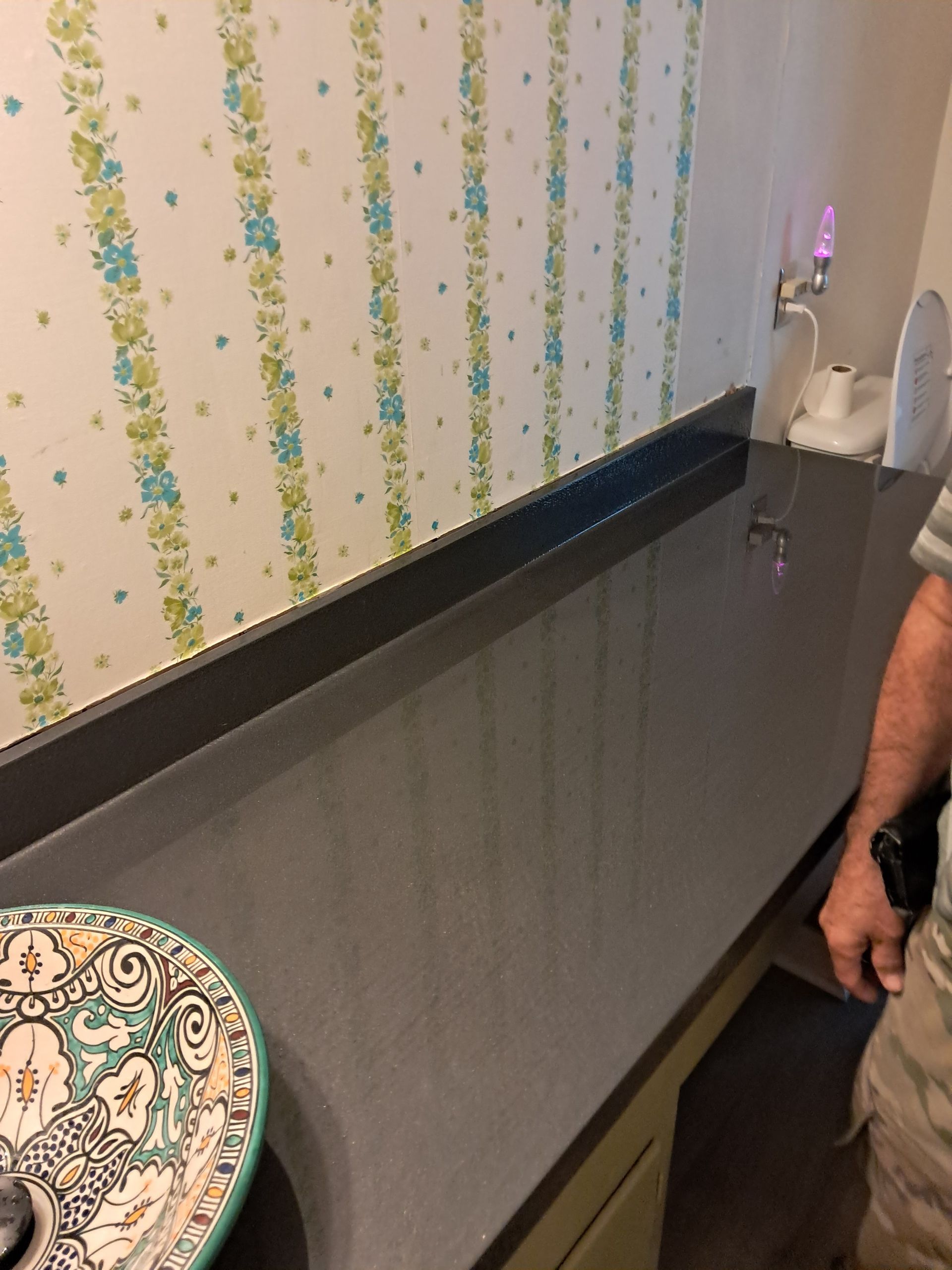 Countertop with patterned wallpaper, a decorative sink, and a person's arm.