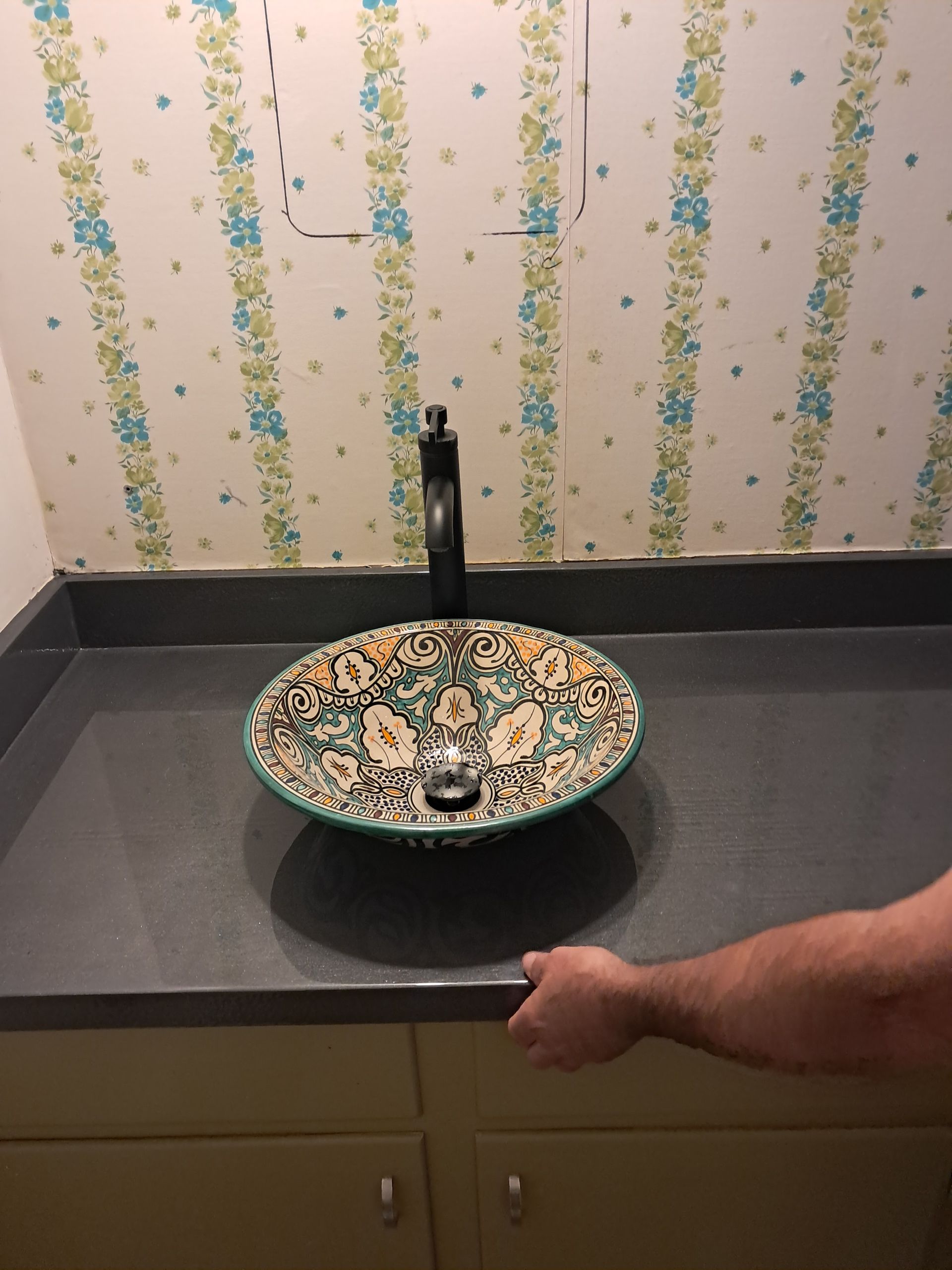 Bathroom with decorative sink and dark faucet on a gray countertop above yellow cabinets; hand reaching.