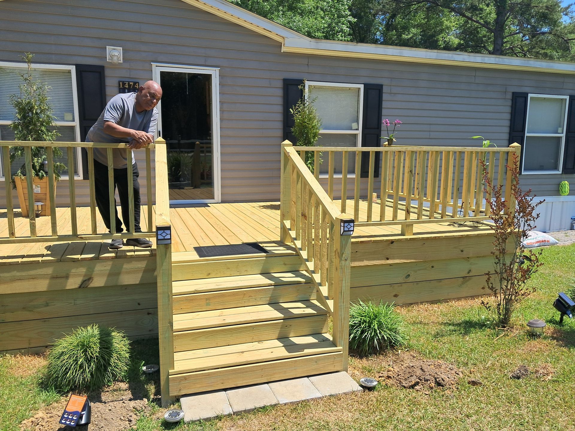 Man leans on a new wooden deck with stairs in front of a gray house. Sunny day.