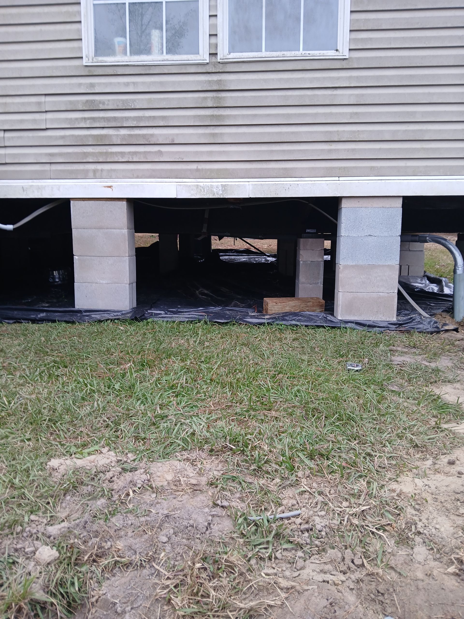Foundation of a house with cinder block supports, dark undercarriage, and grass foreground.