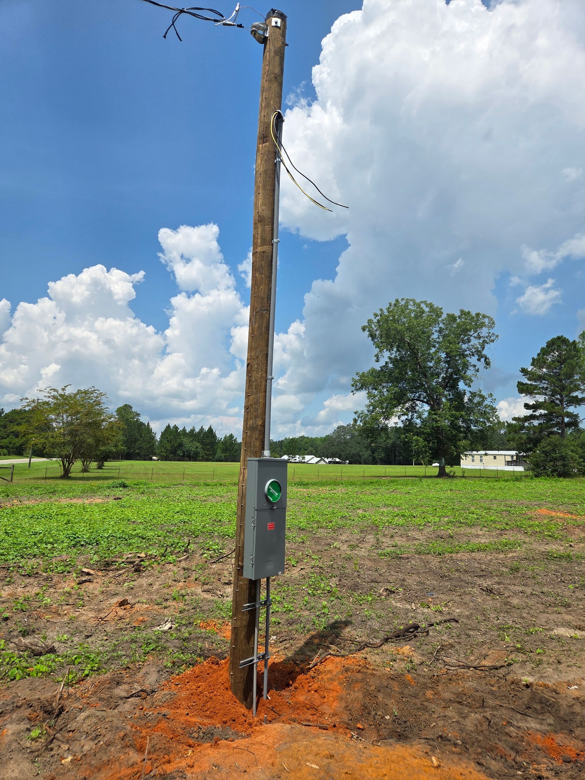 Wooden utility pole with electrical box, in a field, under a cloudy blue sky.