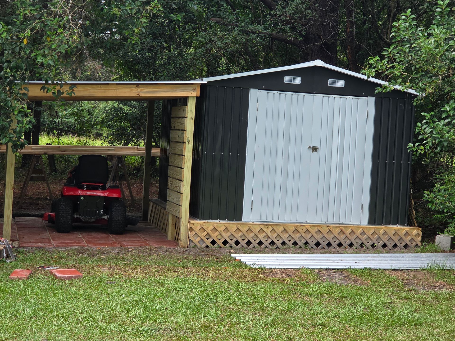 Dark shed with attached carport sheltering a red riding lawnmower on a red-tiled floor.