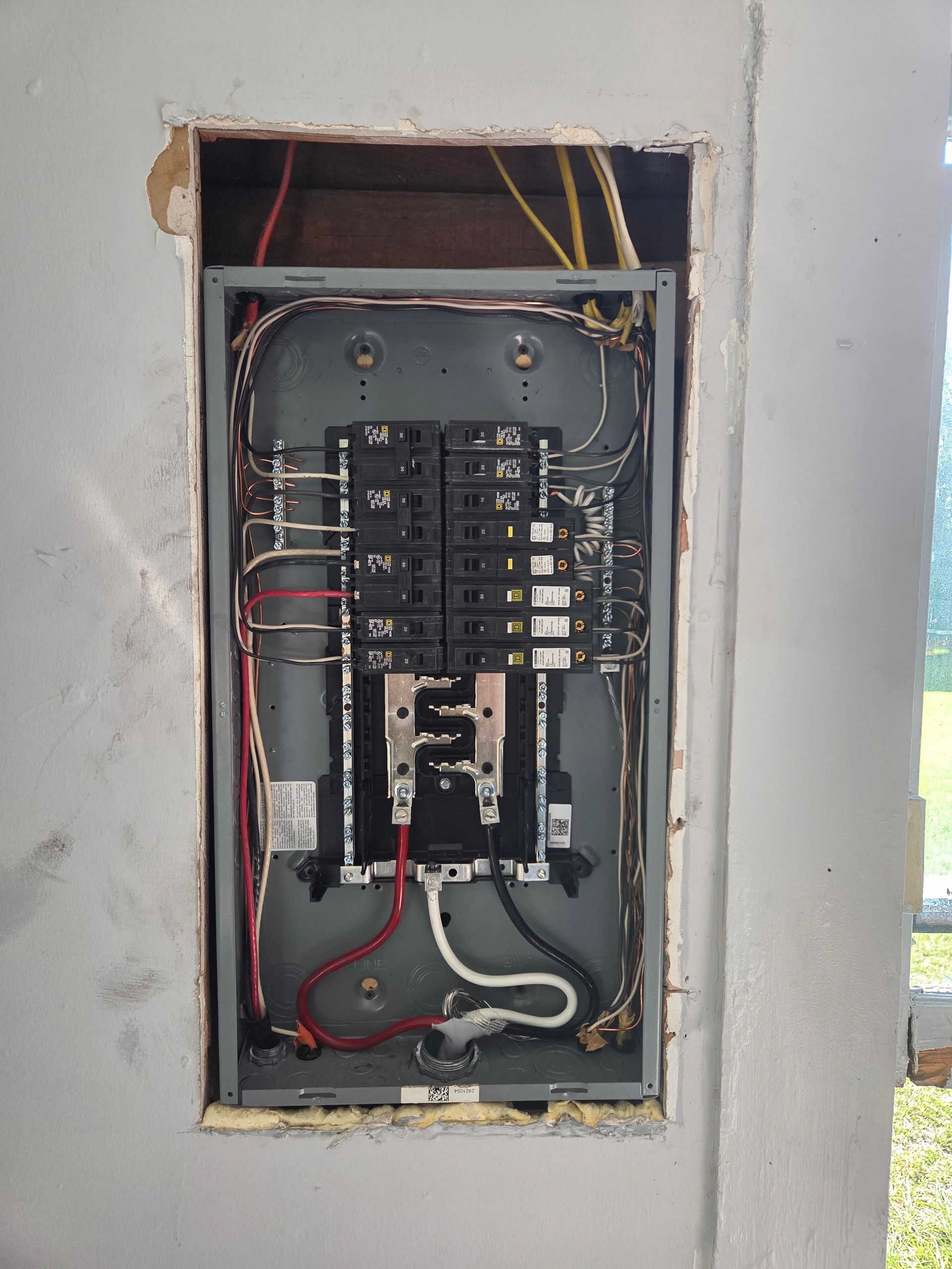 Electrical panel in a gray wall. Black circuit breakers, silver panel, and wires of various colors are visible.