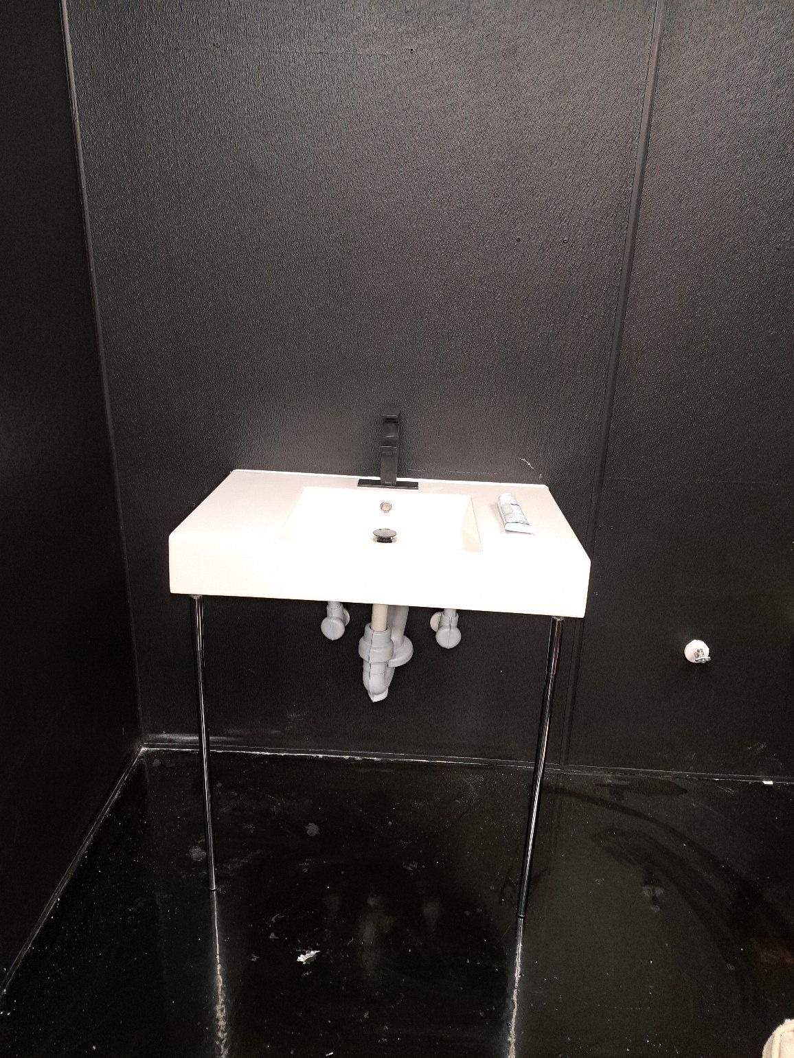 Small white sink mounted on a dark textured wall in a dim restroom stall.