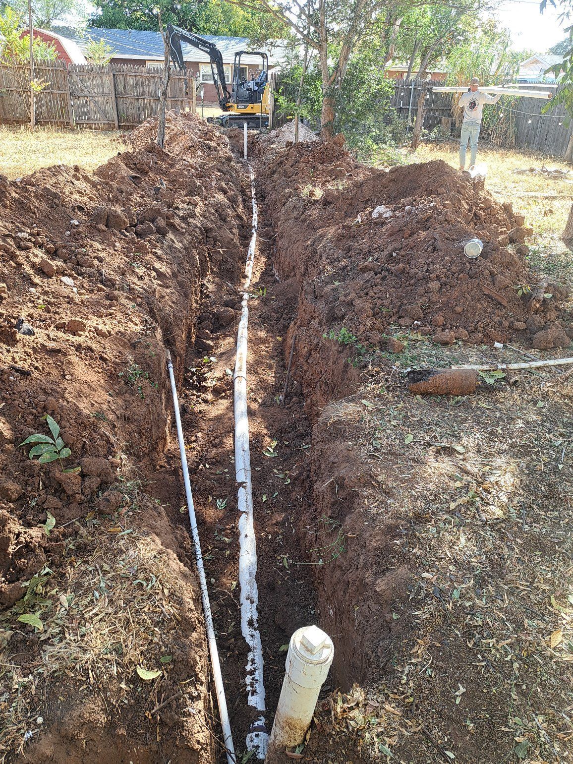Trench with white pipes, backhoe, and a person in a yard. Soil is brown.