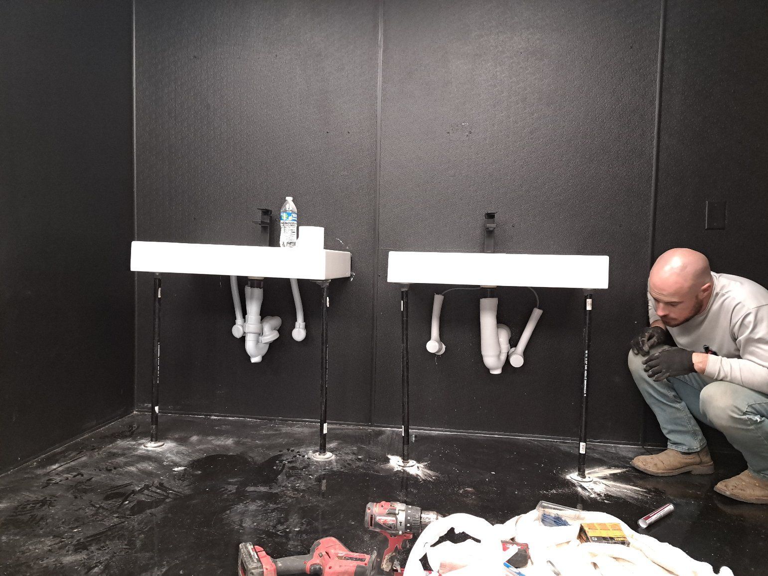 Damaged bathroom with broken sinks and debris on the floor; a man crouches on the right.